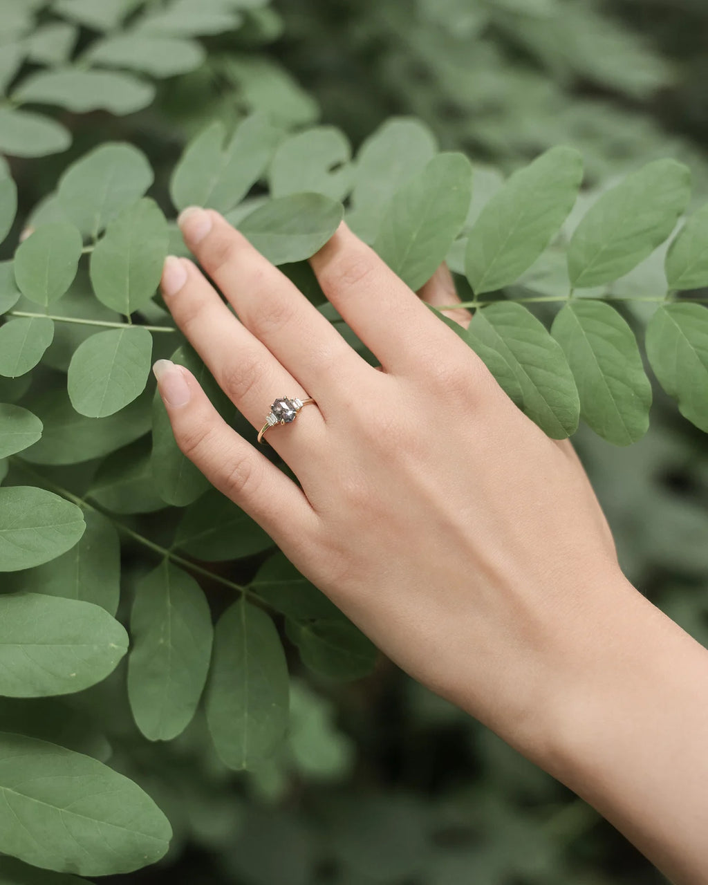 A silver-toned ring with a small clear gemstone worn on a person's ring finger, shown against a background of green leaves.
