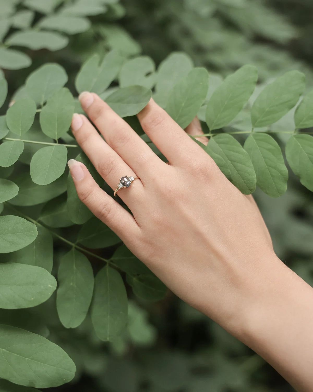 A silver-toned ring with a small clear gemstone worn on a person's ring finger, shown against a background of green leaves.