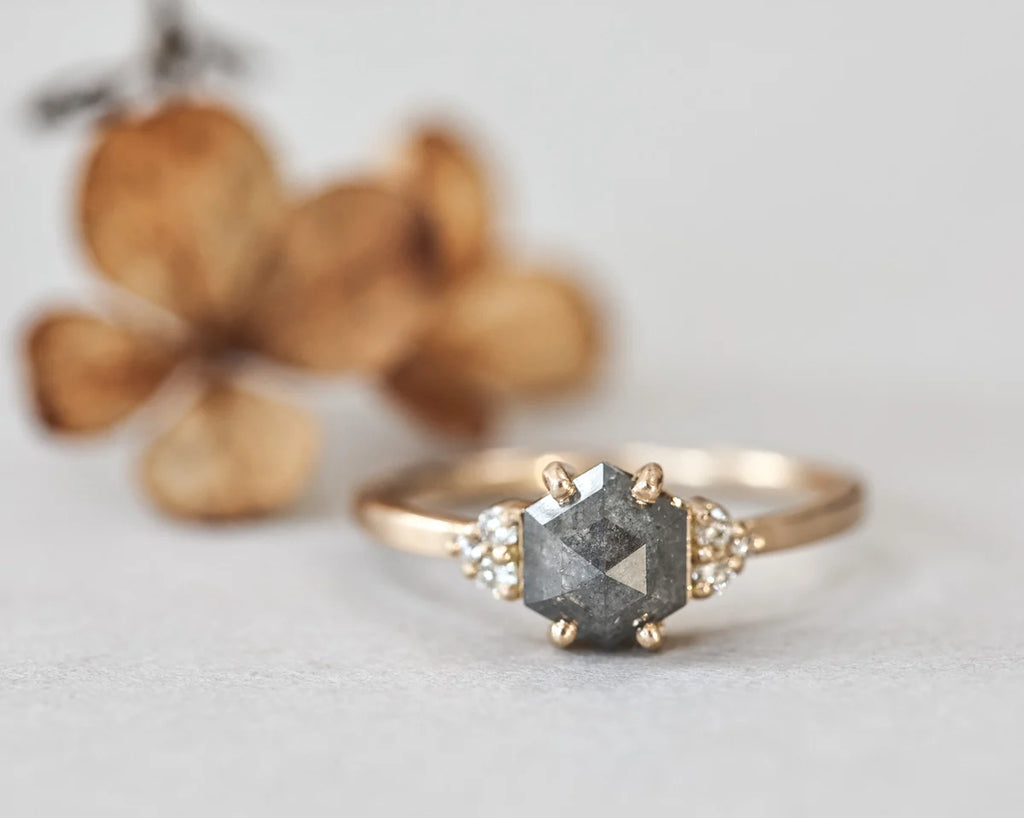 A rose gold ring featuring a large, faceted gray gemstone in a prong setting, accented by small clear stones on either side of the band, photographed against a soft light gray background with blurred dried flowers.