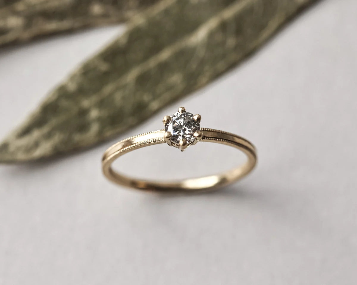A gold solitaire engagement ring with a round-cut clear gemstone, resting on a light gray surface with a blurred olive green leaf in the background.