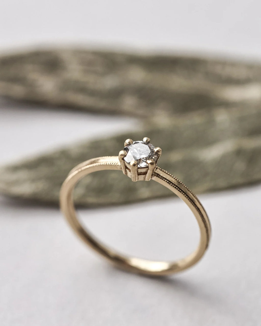 A gold solitaire engagement ring with a round clear gemstone in a six-prong setting, resting on a textured gray stone against a light gray background.