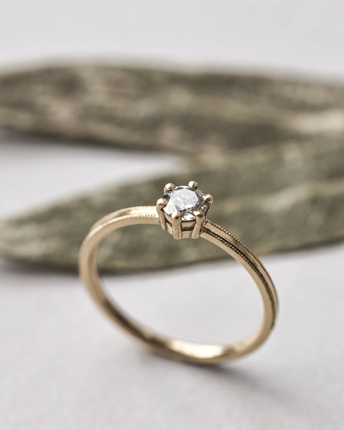 A gold solitaire engagement ring with a round clear gemstone in a six-prong setting, resting on a textured gray stone against a light gray background.