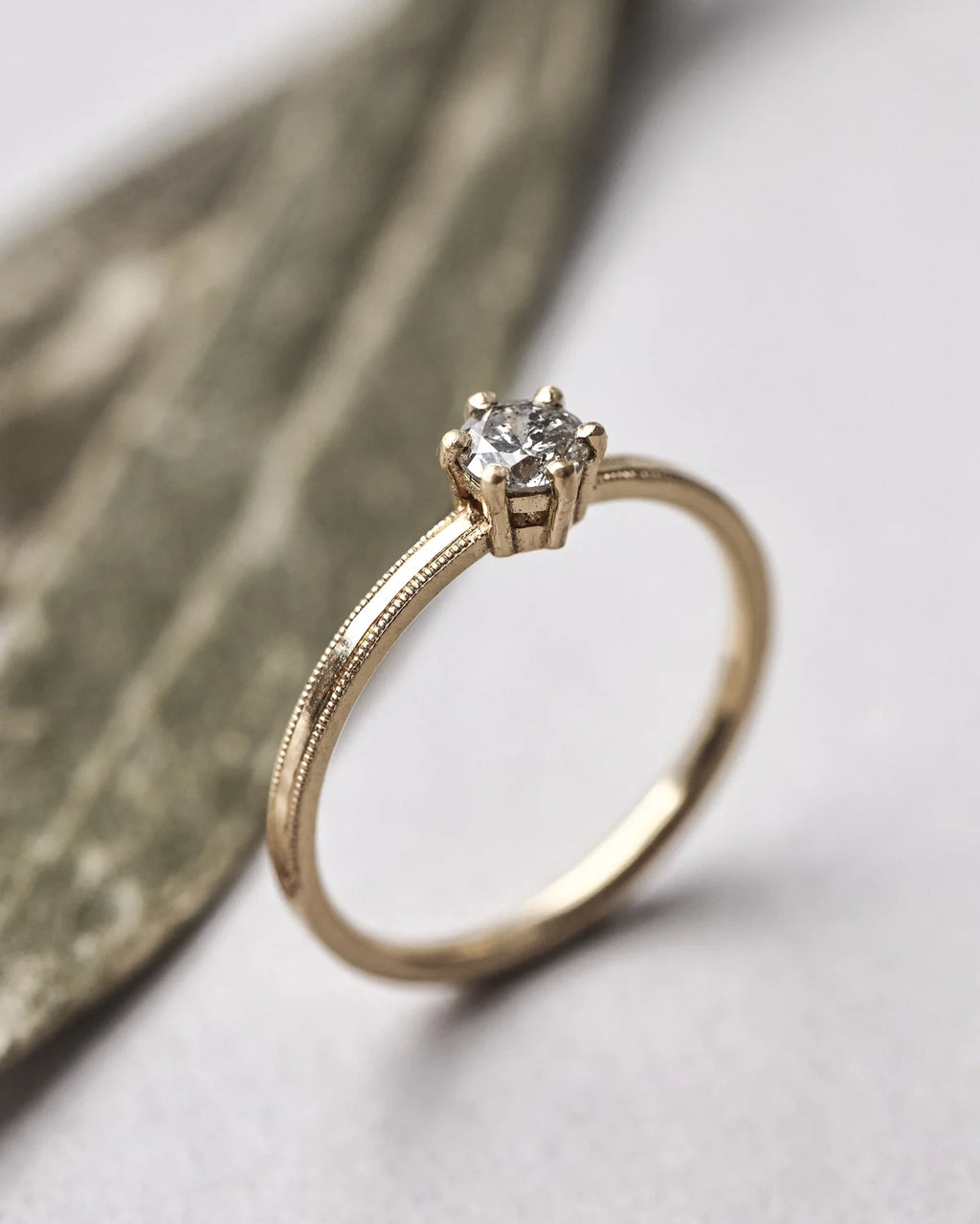 A gold solitaire engagement ring with a round-cut diamond set in a four-prong setting, resting on a textured leaf against a light gray background.