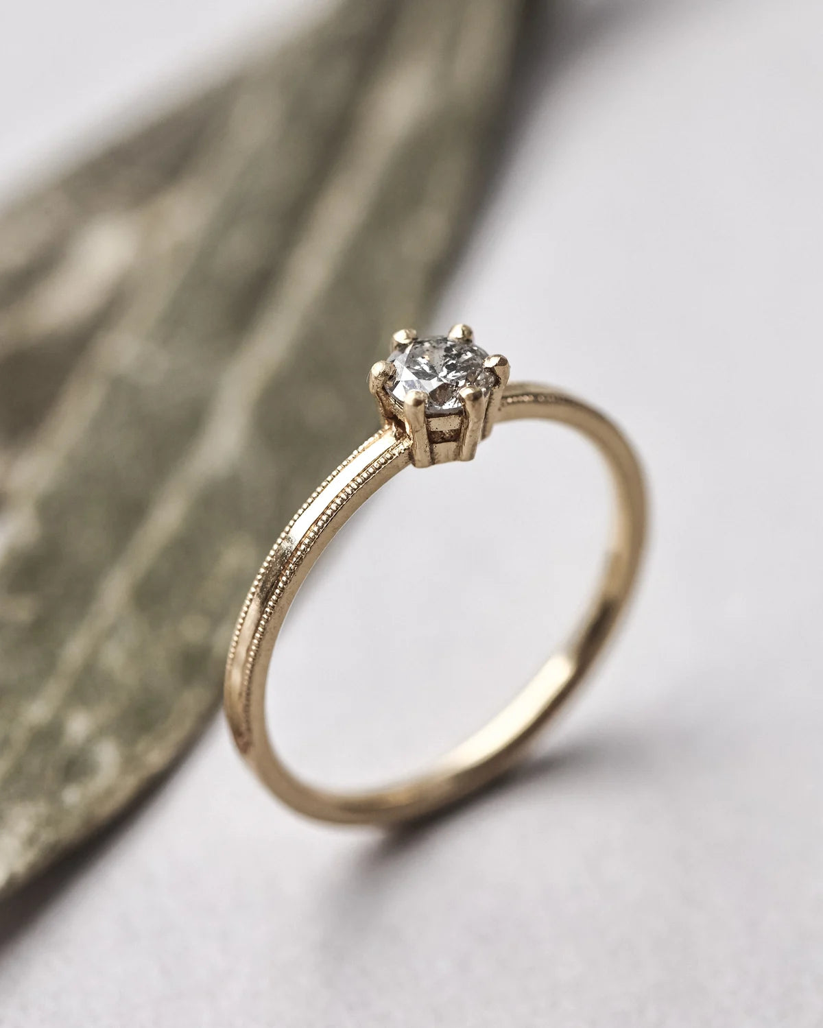 A gold solitaire engagement ring with a round-cut diamond set in a four-prong setting, resting on a textured leaf against a light gray background.