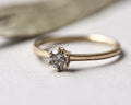 A delicate yellow gold solitaire ring with a single round-cut clear gemstone in a prong setting, resting on a light gray textured surface.