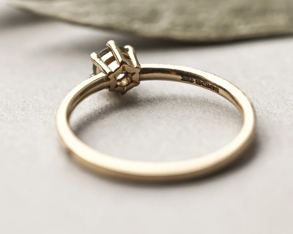 A close-up shot of a yellow gold ring with a four-prong setting, resting on a neutral gray surface with a soft-focus leaf in the background.
