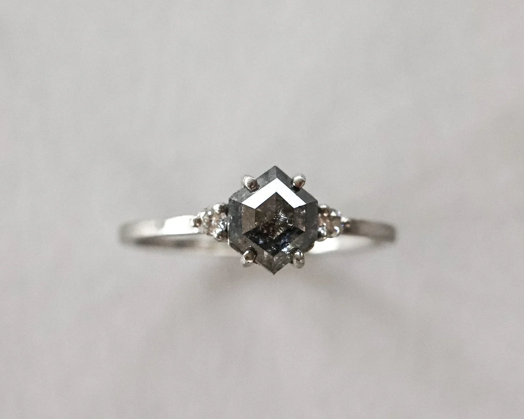 A silver ring featuring a central dark gray gemstone in a square setting, flanked by two small round side stones, photographed against a plain light gray background.
