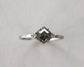 A silver ring featuring a central dark gray gemstone in a square setting, flanked by two small round side stones, photographed against a plain light gray background.