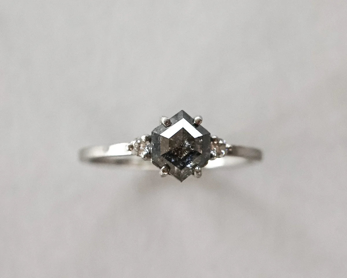 A silver ring featuring a central dark gray gemstone in a square setting, flanked by two small round side stones, photographed against a plain light gray background.