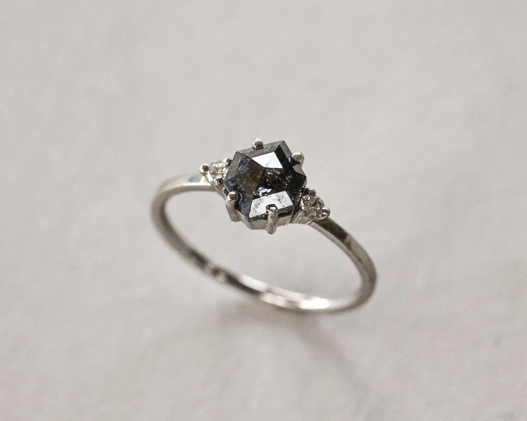 A silver ring featuring a dark gray marquise-cut center stone and small clear accent stones on either side, set against a neutral light gray background.