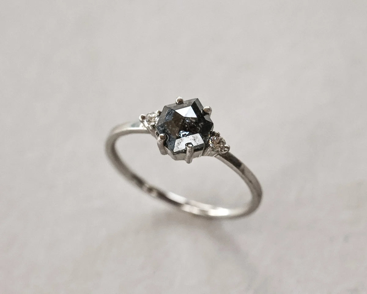 A silver ring featuring a dark gray marquise-cut center stone and small clear accent stones on either side, set against a neutral light gray background.