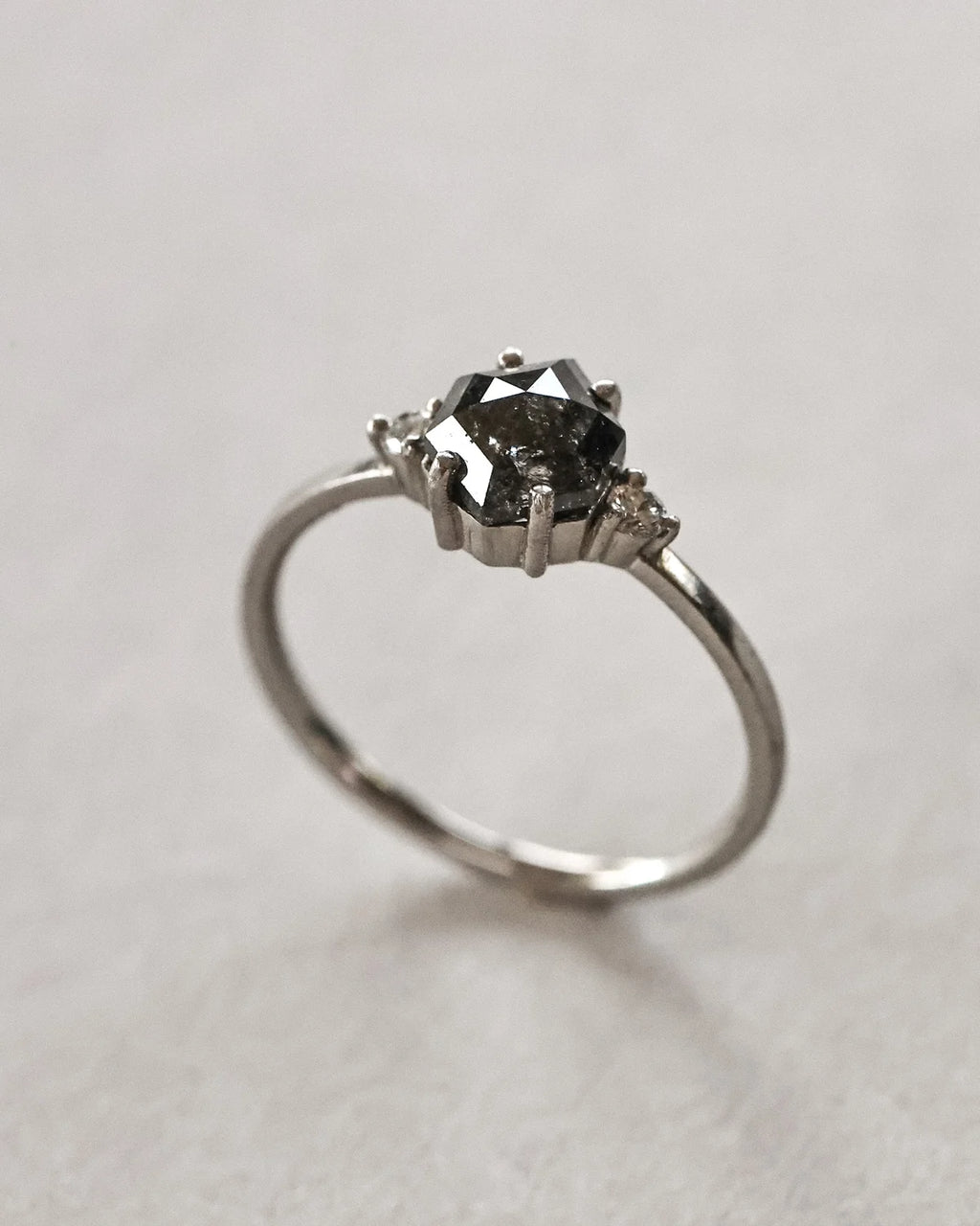 A silver-toned ring featuring a dark, faceted round center stone held in a prong setting, with small side stones, photographed on a neutral light gray background.