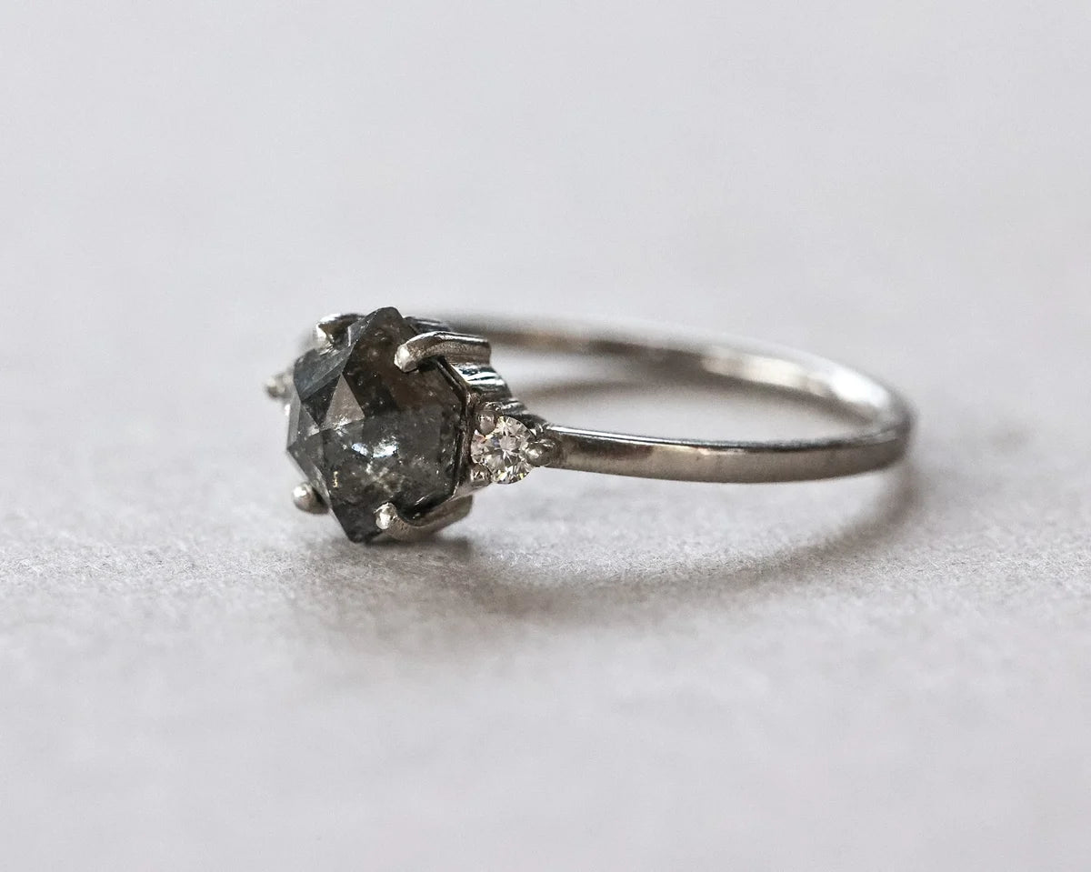 A silver ring featuring a large, rough-cut dark gray gemstone in a prong setting, accented by small clear stones on the band, photographed on a neutral light gray background.