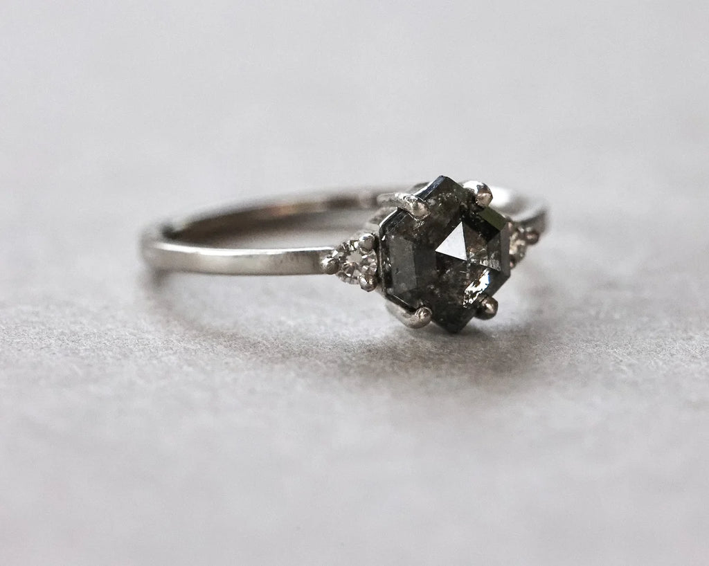 A silver-toned ring featuring a central dark gray faceted gemstone in a prong setting, accented by small clear stones on either side, photographed on a light gray textured surface.