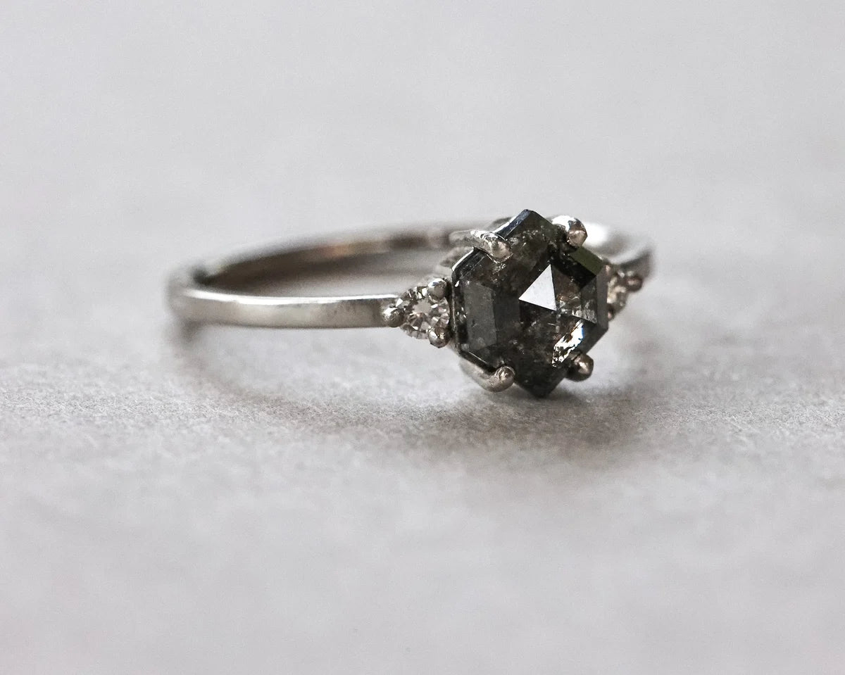A silver-toned ring featuring a central dark gray faceted gemstone in a prong setting, accented by small clear stones on either side, photographed on a light gray textured surface.