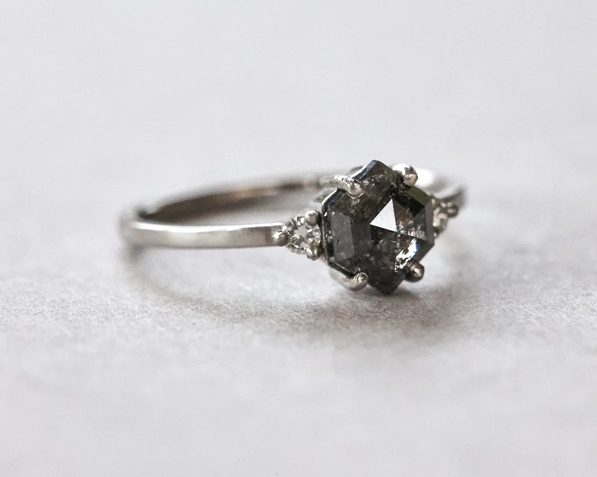 A silver ring featuring a central dark gray faceted gemstone and two small clear accent stones on either side, photographed on a soft light gray background.