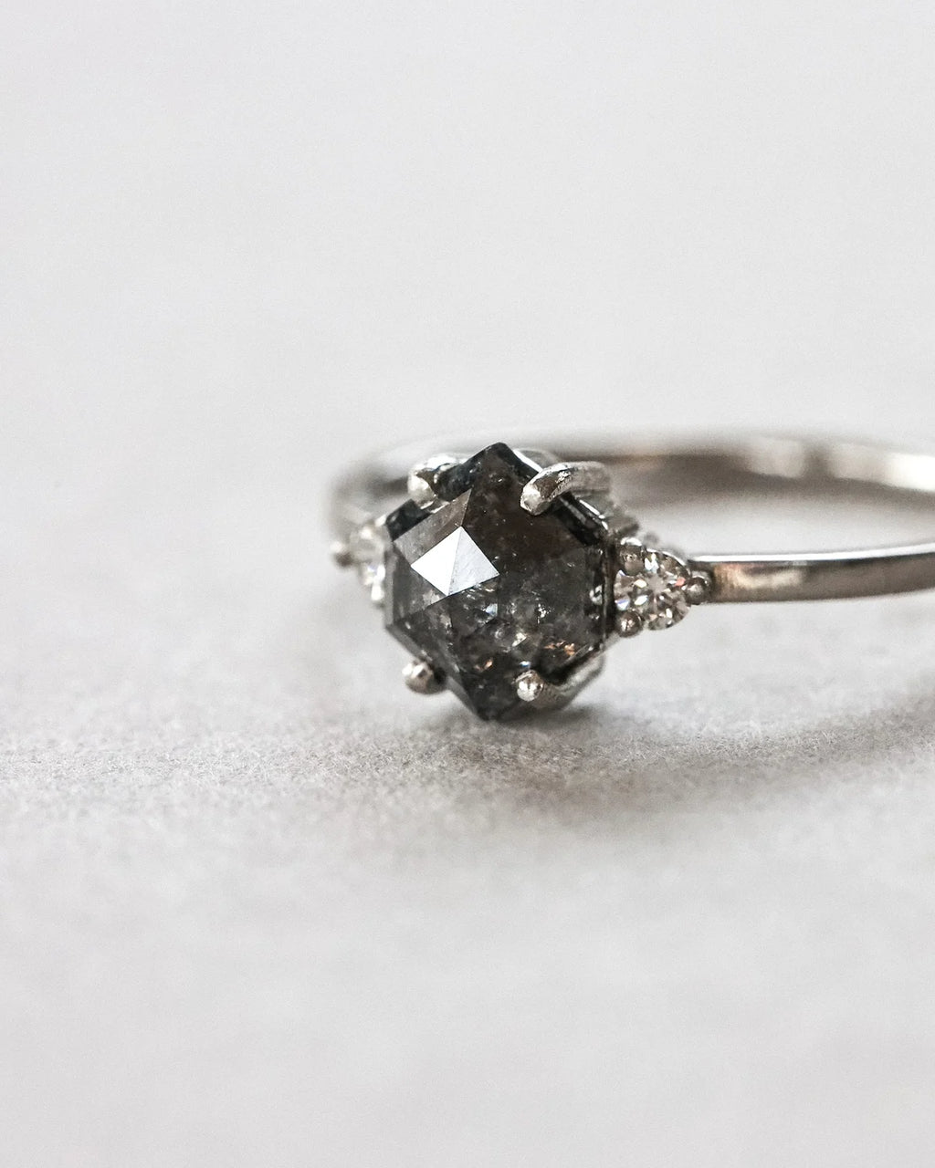 A close-up shot of a silver ring featuring a dark gray, faceted cushion-cut center stone and small clear accent stones on either side of the band, set against a soft, light gray background.
