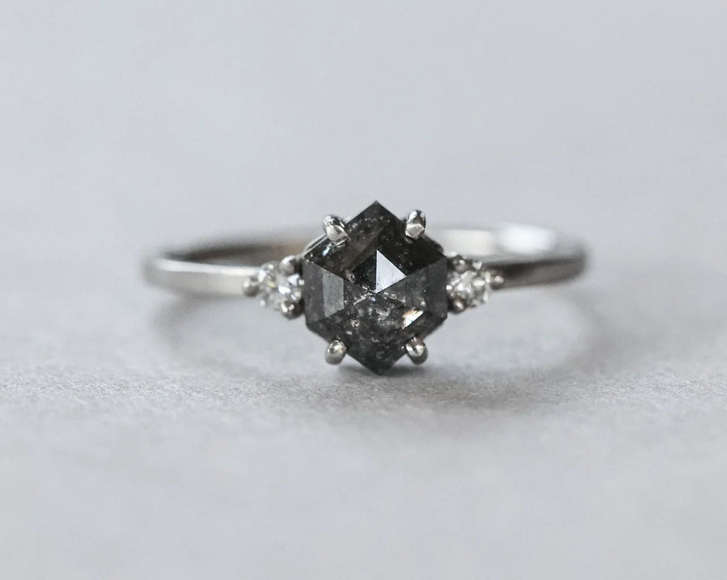 A silver-toned ring featuring a central dark gray marquise-cut gemstone in a prong setting, accented by two small clear round-cut stones on either side of the band, photographed on a neutral light gray background.