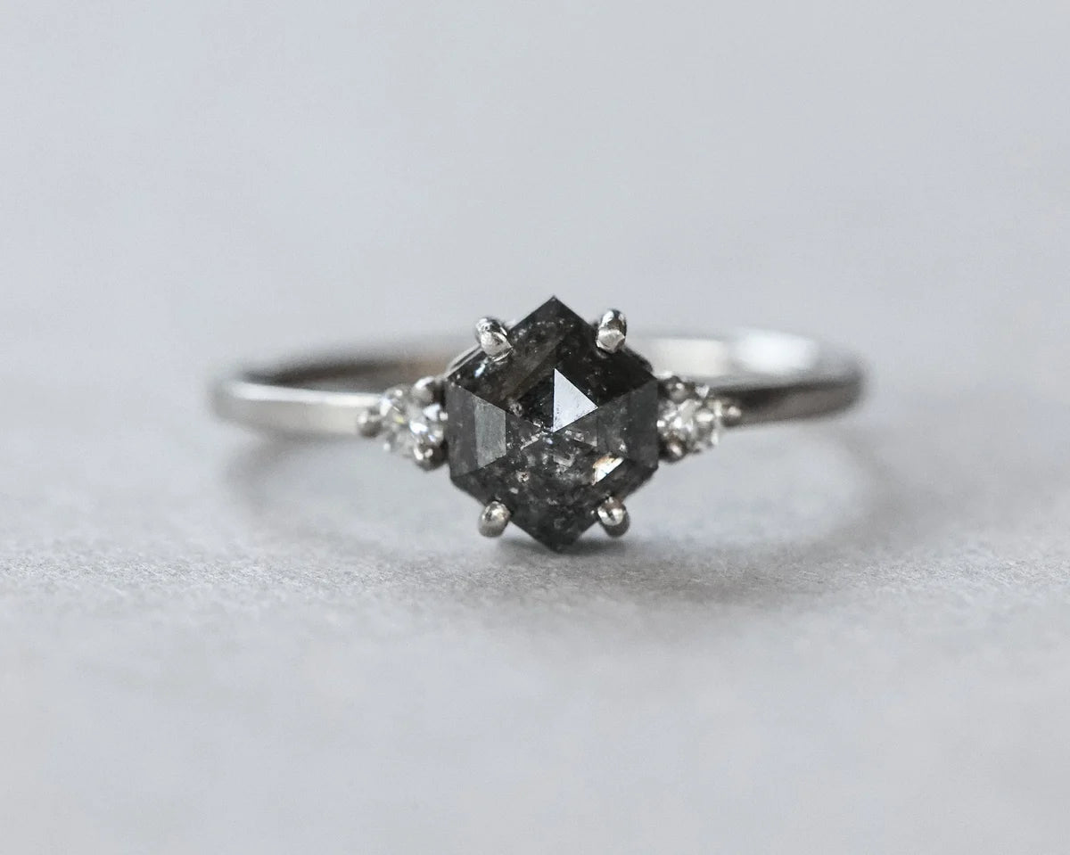 A silver-toned ring featuring a central dark gray marquise-cut gemstone in a prong setting, accented by two small clear round-cut stones on either side of the band, photographed on a neutral light gray background.