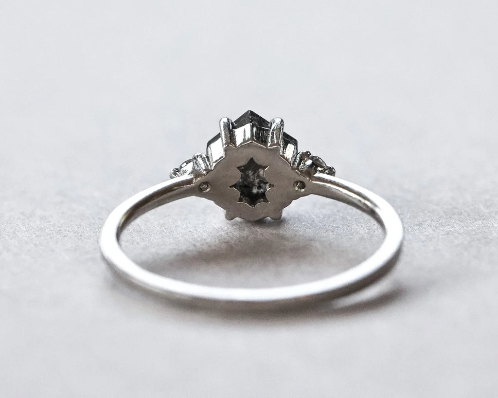 A silver ring with a geometric starburst cluster setting featuring a central black gemstone and small accent stones, shown on a neutral light gray background.