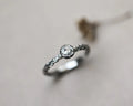 A silver-toned twisted band ring with a round clear gemstone, photographed on a soft light gray background with a blurred dried flower prop.