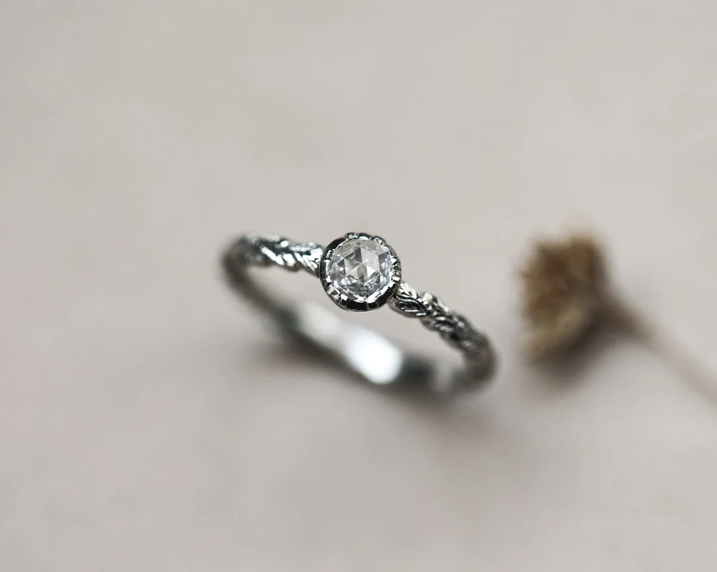 A silver-toned ring with a round clear gemstone and a textured twisted band, photographed on a soft light beige background with a blurred dried flower prop.