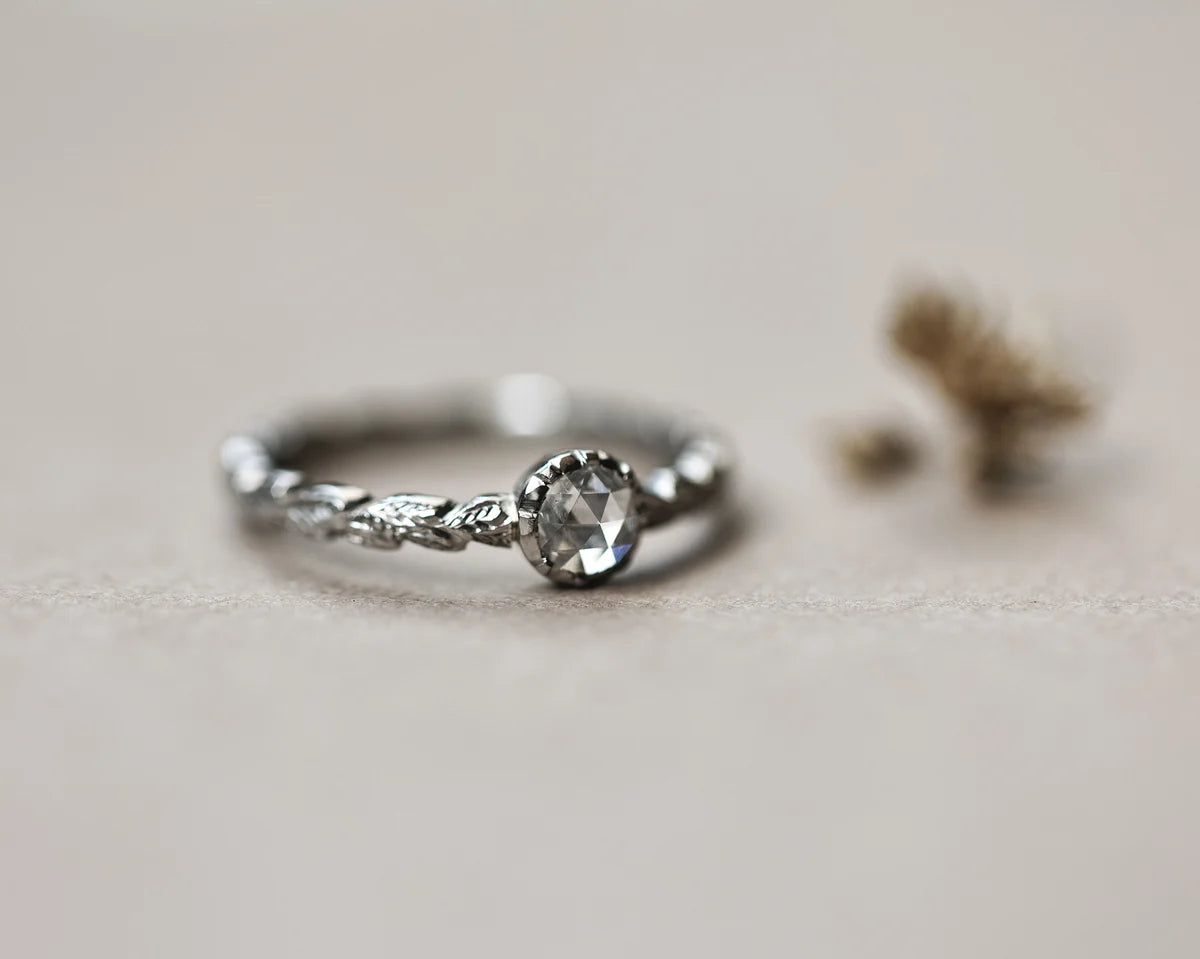 A platinum ring with a carved band and a central clear faceted diamond, photographed on a soft beige surface with a blurred decorative element in the background.