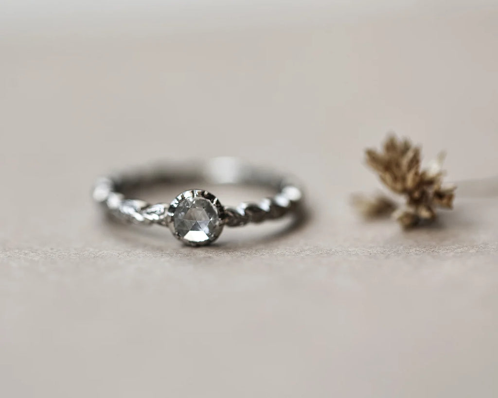 A Platinum ring with a twisted band and a central clear gemstone, photographed on a soft beige surface with a small dried flower prop at the side.