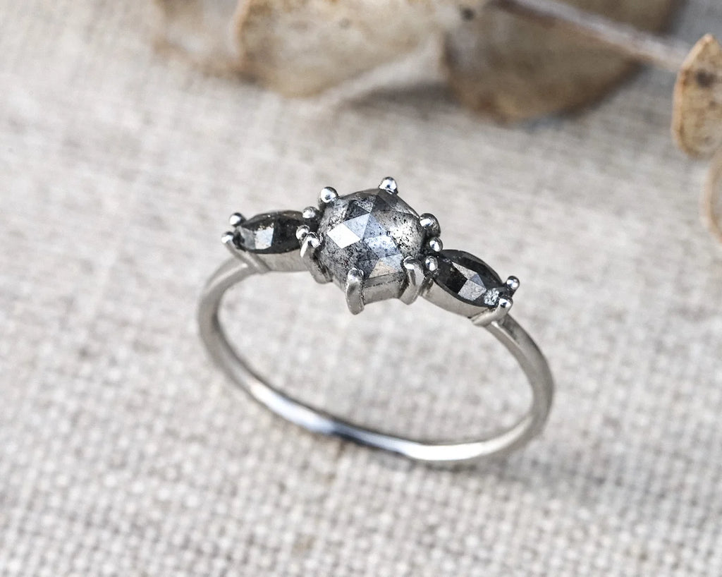 A silver ring featuring a central round gray gemstone flanked by two smaller marquise-shaped gray gemstones, resting on a textured beige fabric background.