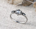 A silver ring featuring a central round gray gemstone flanked by two smaller marquise-shaped gray gemstones, resting on a textured beige fabric background.