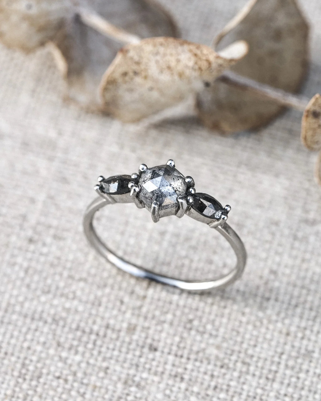 A silver ring featuring a central round clear gemstone flanked by two small marquise-cut dark gray gemstones, resting on a textured beige fabric with dried botanical elements in the background.
