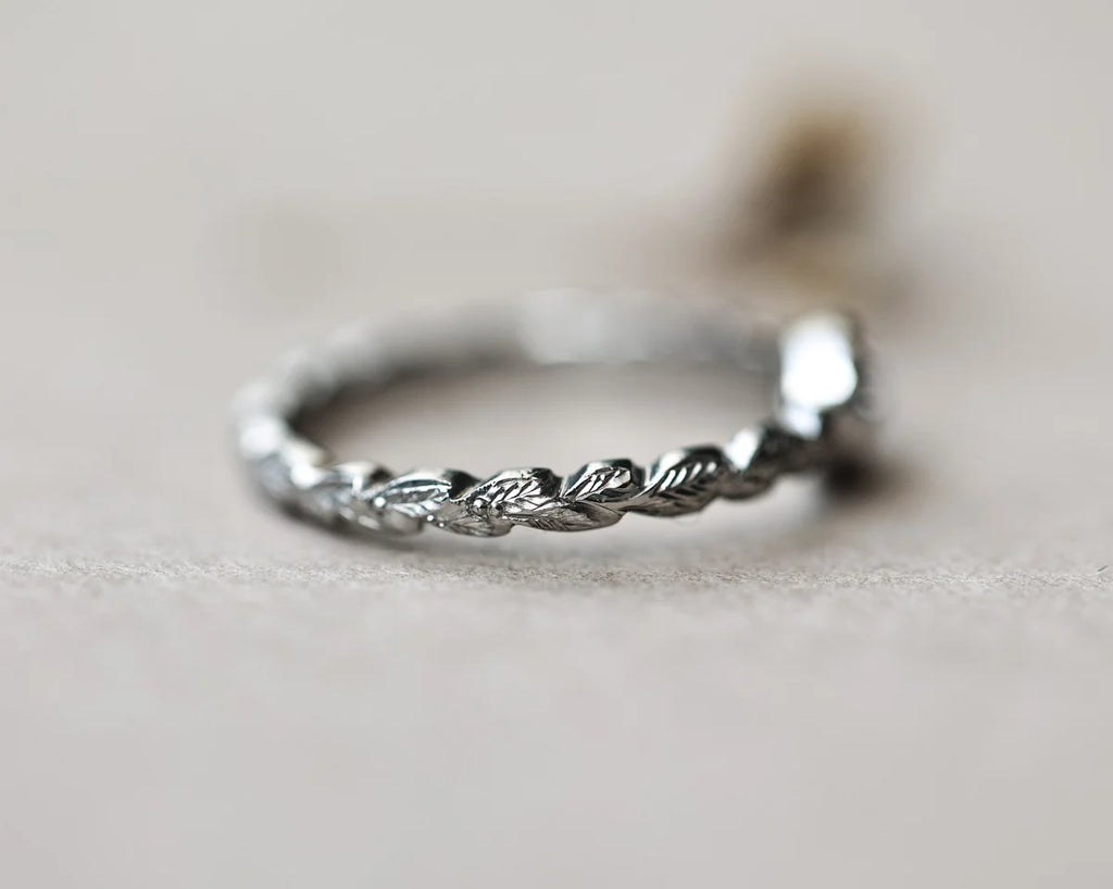 A platinum twisted rope band ring with a small clear gemstone, photographed in a close-up macro shot on a neutral beige background.