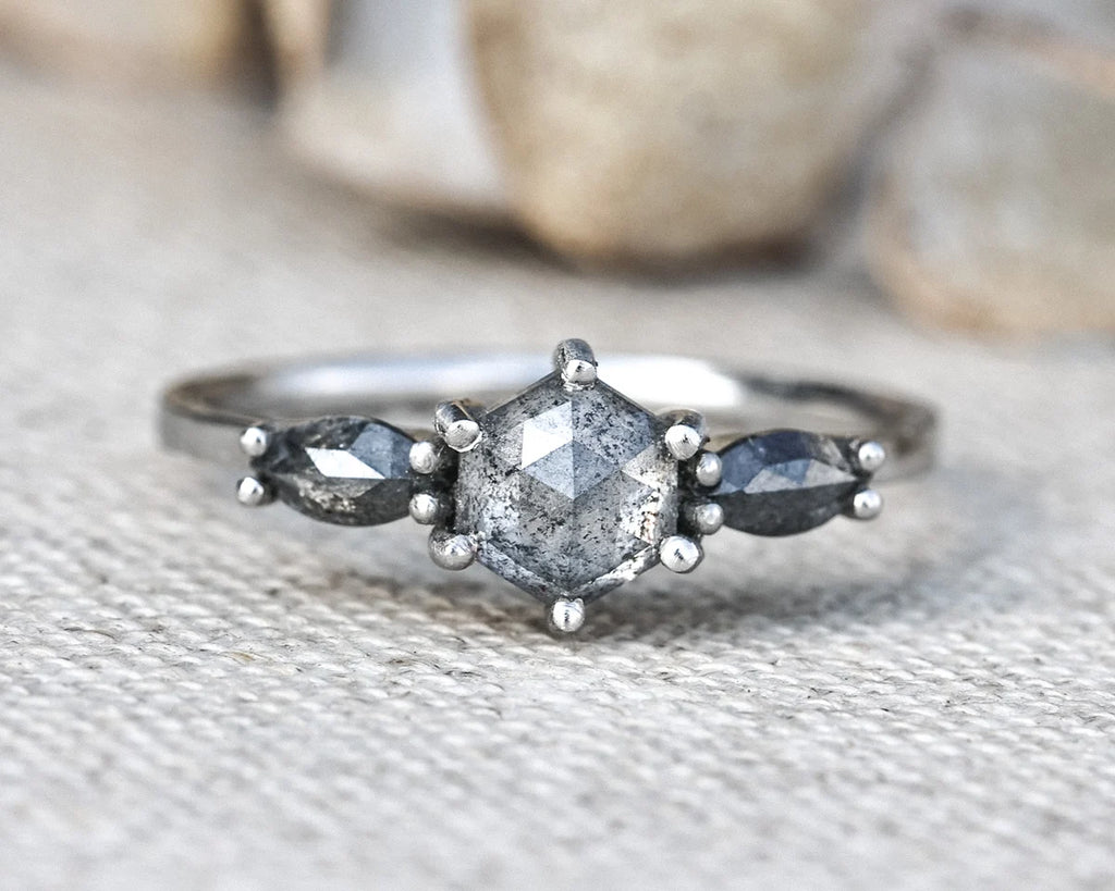 A silver ring featuring a central round gray gemstone flanked by two marquise-shaped dark gray stones, resting on a textured beige fabric surface with a blurred natural background.