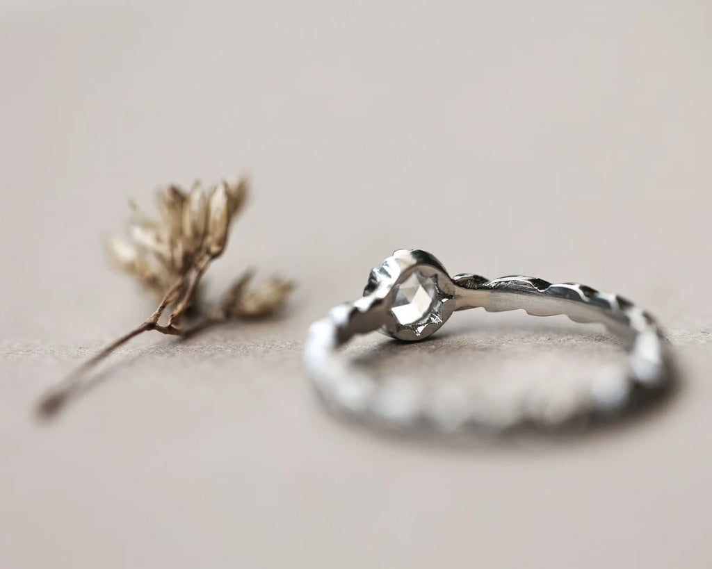 A platinum ring with a textured, hammered band and a heart-shaped clear gemstone, displayed on a neutral beige surface next to a small dried flower sprig.