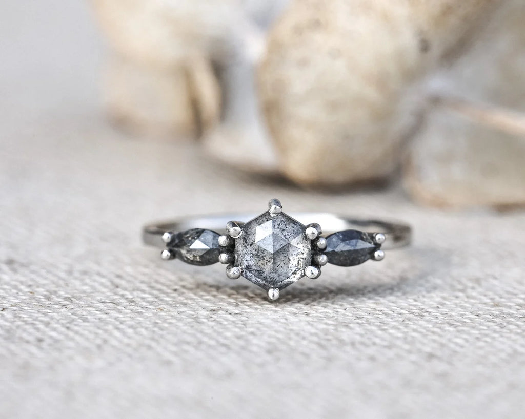 A silver ring featuring a central clear faceted gemstone flanked by two small dark gray marquise-cut gemstones, resting on a textured beige fabric surface with blurred stones in the background.
