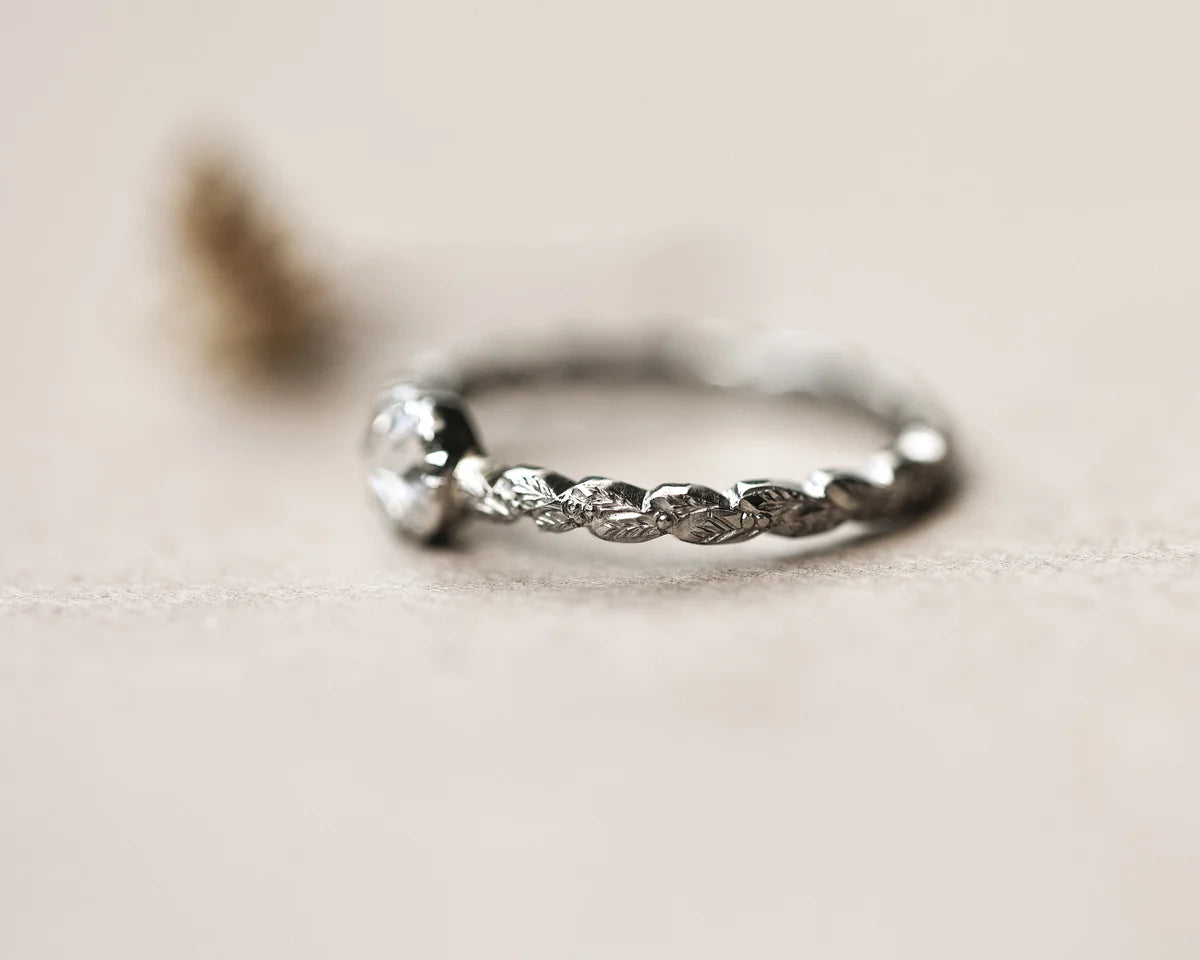 A platinum ring with a carved textured, leaf-like twisted band and a single clear gemstone, photographed on a soft beige background.