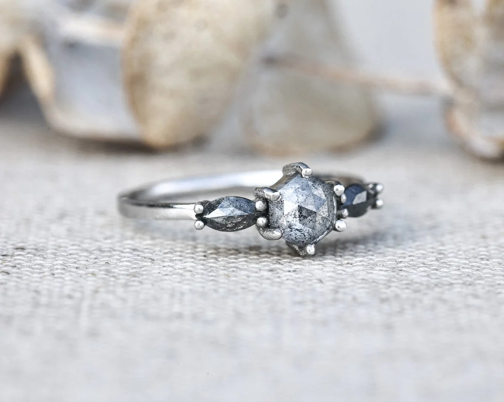 A silver ring featuring a central round clear gemstone flanked by two marquise-cut dark gray stones and small round dark blue stones, resting on a textured light gray surface with blurred decorative stones in the background.