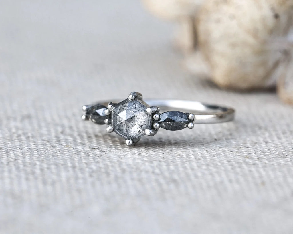 A silver ring featuring a central clear faceted gemstone surrounded by small metallic beads, with two dark gray marquise-shaped side stones, photographed on a textured light fabric with a blurred shell in the background.