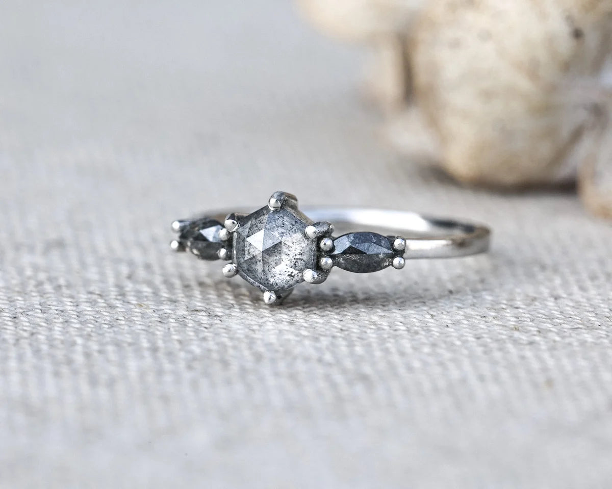 A silver ring featuring a central clear faceted gemstone surrounded by small metallic beads, with two dark gray marquise-shaped side stones, photographed on a textured light fabric with a blurred shell in the background.
