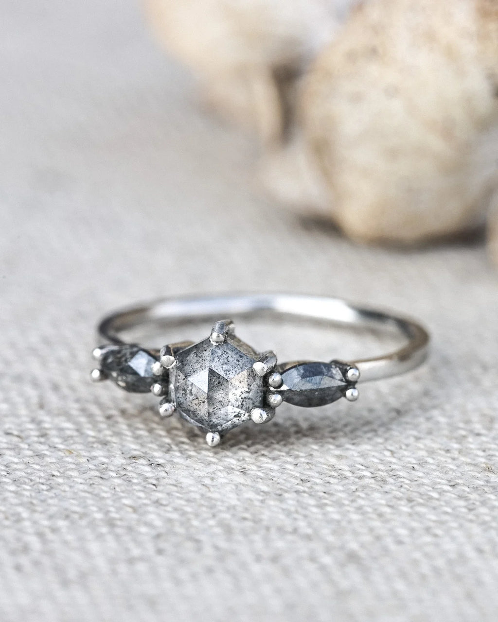 A silver ring featuring a central faceted gray gemstone and two smaller marquise-shaped gray gemstones on either side, resting on a textured beige fabric surface with a blurred natural background.