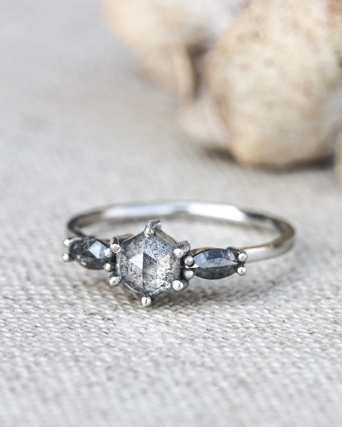 A silver ring featuring a central faceted gray gemstone and two smaller marquise-shaped gray gemstones on either side, resting on a textured beige fabric surface with a blurred natural background.