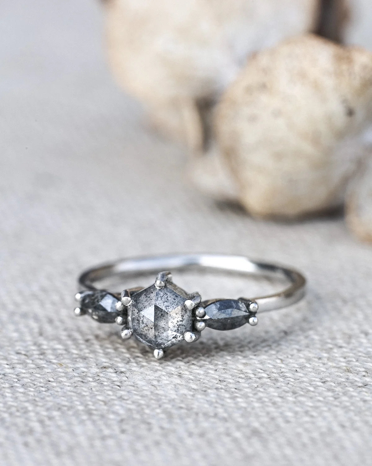 A silver ring featuring a central round gray gemstone flanked by two smaller marquise-shaped gray stones, resting on a textured beige fabric with blurred stones in the background.