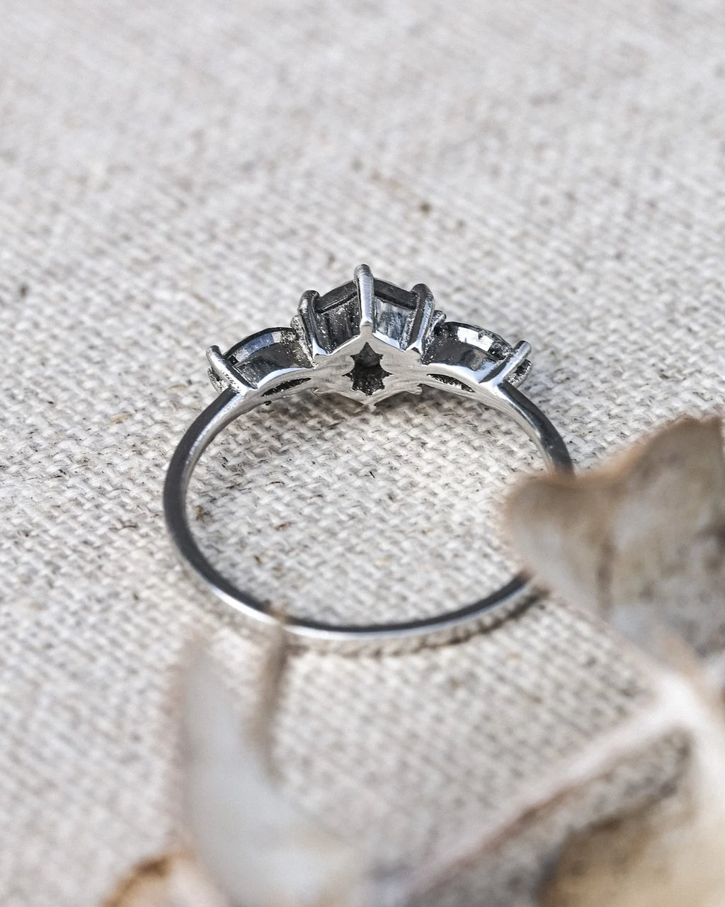A silver ring with a star-shaped halo setting, photographed close-up on a textured beige fabric background with blurred dried leaves in the foreground.