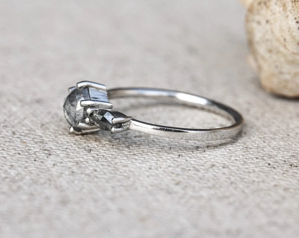A silver ring with a unique multi-prong setting holding three dark gray stones, resting on a textured beige fabric surface with a blurred stone in the background.