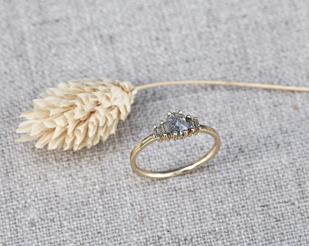 A gold ring with a small gray gemstone cluster setting, displayed on a textured gray fabric next to a dried beige flower.