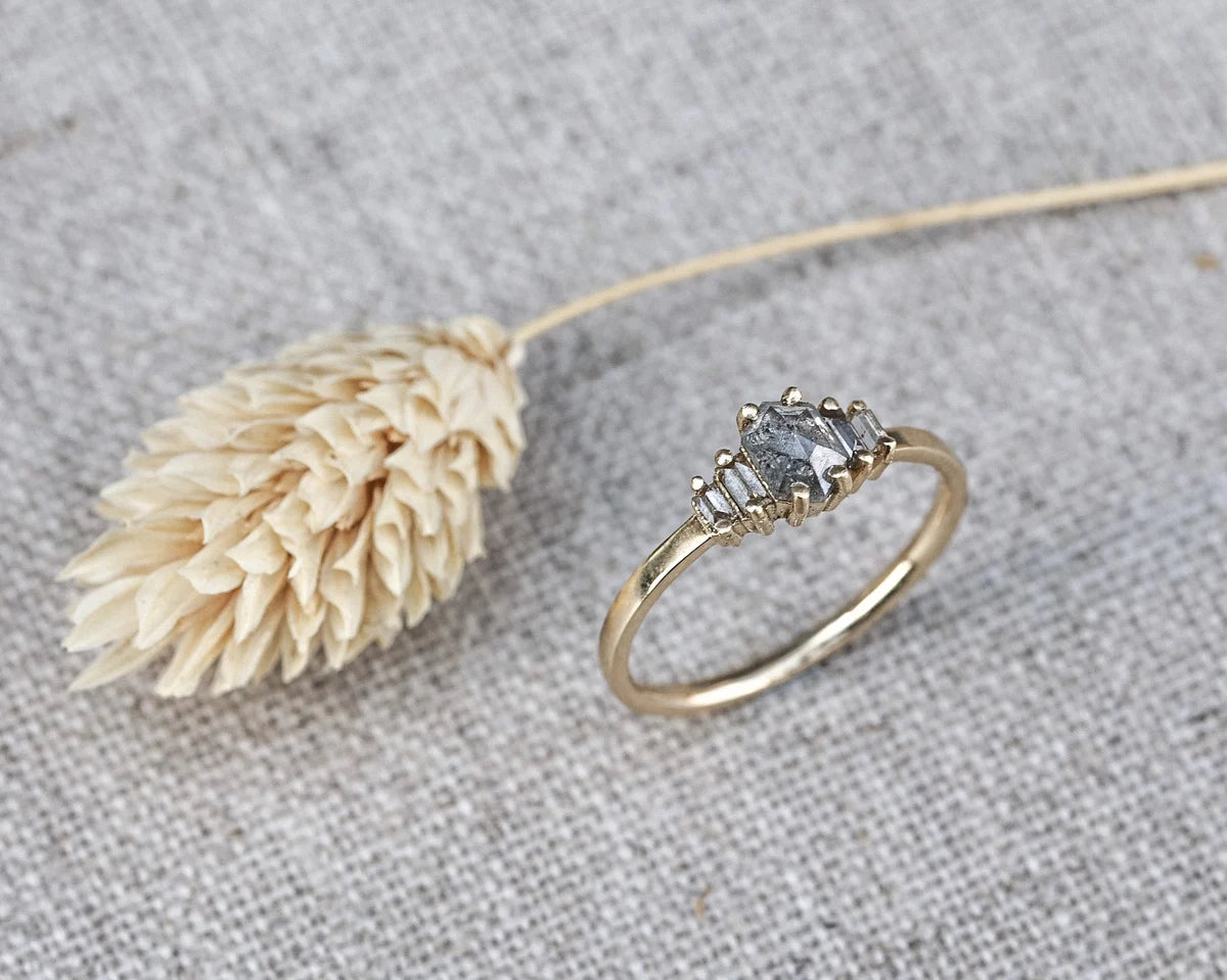 A gold ring with a three-stone cluster setting featuring gray-blue stones, styled on a textured gray linen background next to a dried beige flower.