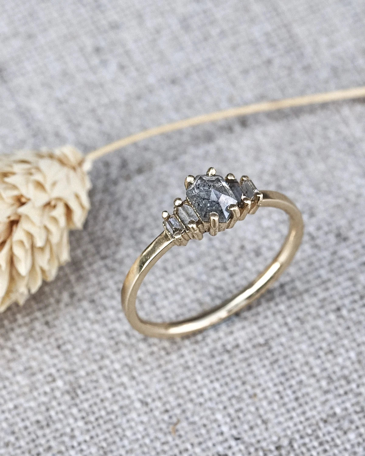 A gold ring with a three-stone cluster featuring a central gray gemstone and two smaller clear stones, displayed on a textured gray fabric next to a dried flower.