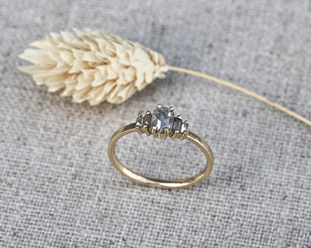 A gold ring with a three-stone cluster featuring a central round blue gemstone and two smaller marquise-cut stones, displayed on a textured gray linen background next to a dried wheat stalk.
