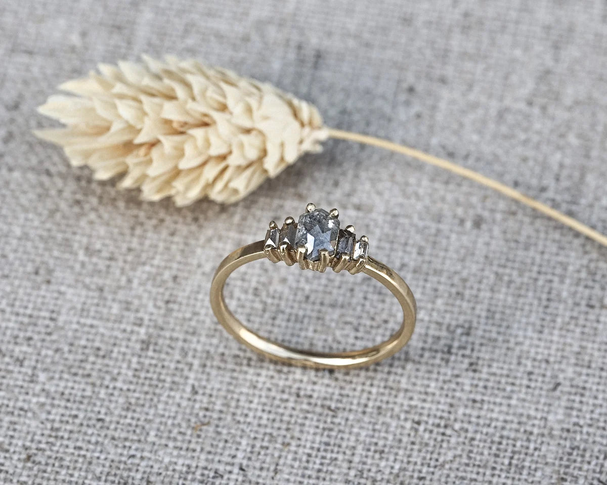 A gold ring with a three-stone cluster featuring a central round blue gemstone and two smaller marquise-cut stones, displayed on a textured gray linen background next to a dried wheat stalk.