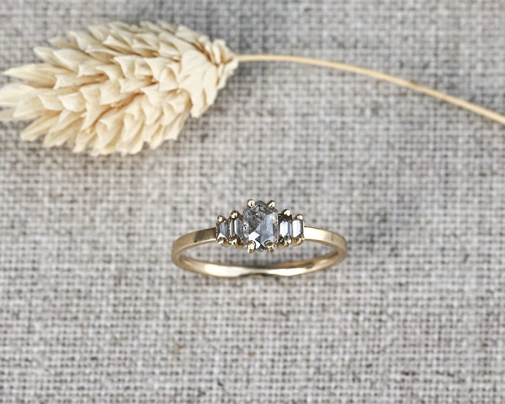 A gold ring with three marquise-cut clear gemstones, displayed on a textured beige fabric background with a dried wheat stalk prop.